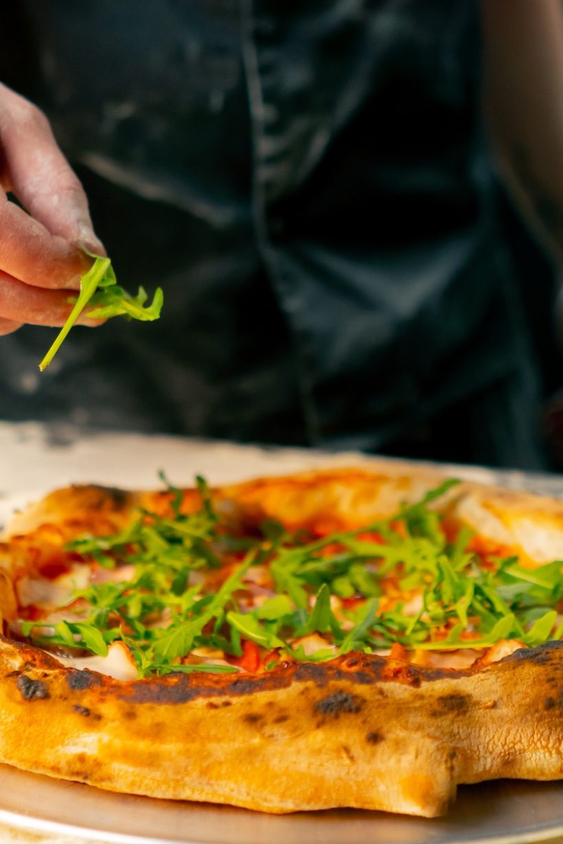 close up in a professional kitchen in a pizzeria a young chef in a black uniform puts ingredients on pizza